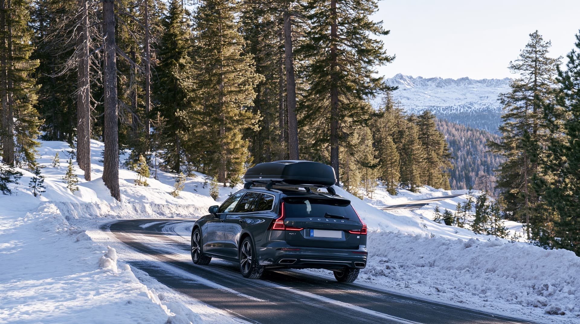 An estate car with a roof box on a winding mountain road through snowy pine forest, with sunlit peaks ahead